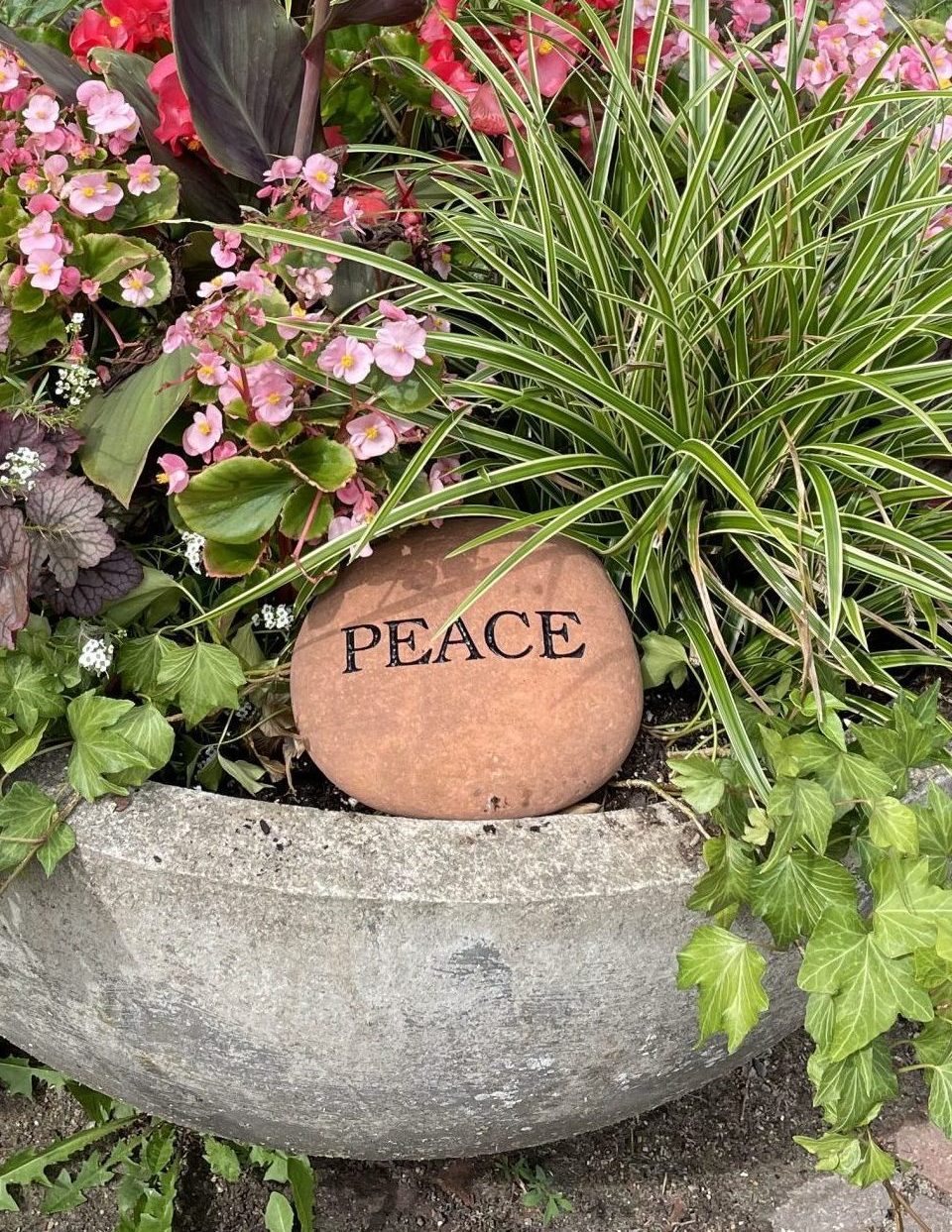 A large stone pot filled with green and pink flowers and plants. A smooth, round stone with the word "PEACE" is placed at the front. The pot is set on a stone pathway amidst grass and a large sculpture in the background.