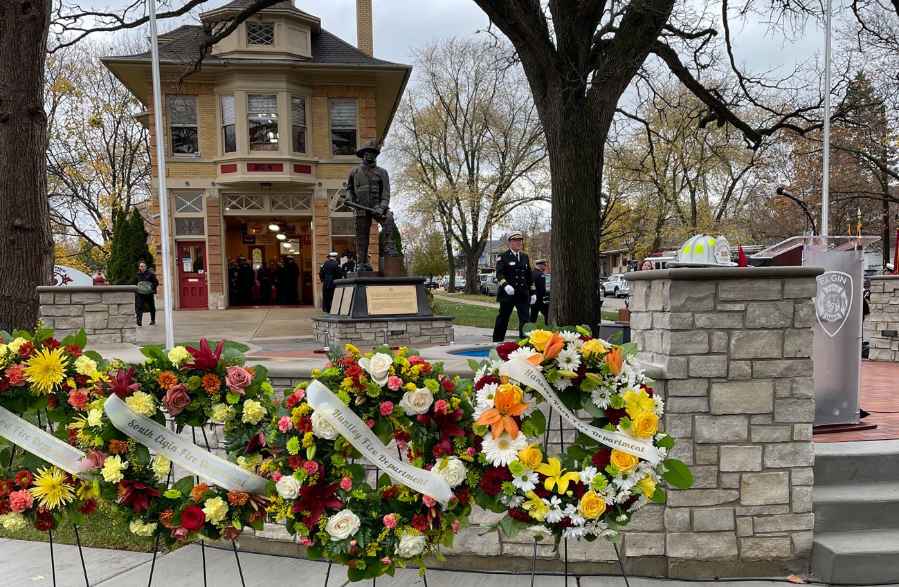 A memorial site with a statue and several wreaths adorned with flowers and ribbons. Behind the stone wall is a two-story building with people gathered. Trees with autumn leaves frame the scene, and a man in uniform stands near the statue.