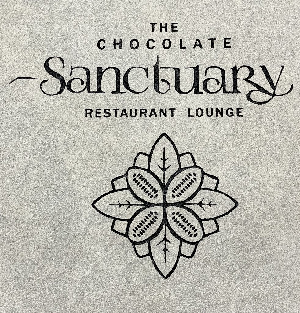 Text reading “The Chocolate Sanctuary Restaurant Lounge” is above a stylized floral design resembling cacao pods and leaves. The background is a textured gray surface.