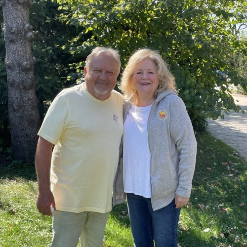 A man in a pale yellow shirt and light pants stands next to a woman in a gray jacket and jeans. They are outdoors on a sunny day, with trees and grass in the background. Both are smiling at the camera.