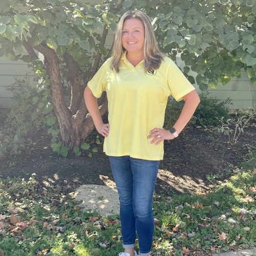 A woman stands smiling in front of a tree, wearing a yellow shirt, blue jeans, and white sneakers. She has long hair and her hands on her hips. Sunlight filters through the leaves, casting dappled shadows on the ground.