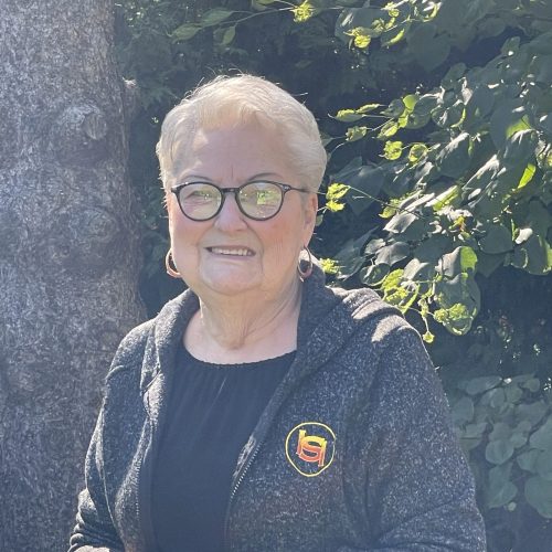 Elderly person with short white hair and glasses, smiling in a shaded area. Wearing a dark jacket with a yellow and red logo, standing in front of lush green foliage and a tree trunk. Sunlight creates a dappled effect on the scene.