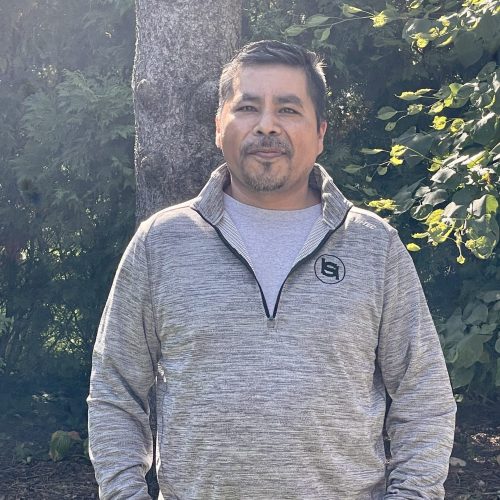 A person stands outdoors in front of a tree and greenery, wearing a gray quarter-zip sweater and a T-shirt. They have short dark hair and facial hair, and they are smiling slightly at the camera. The sunlight creates a soft glow in the background.